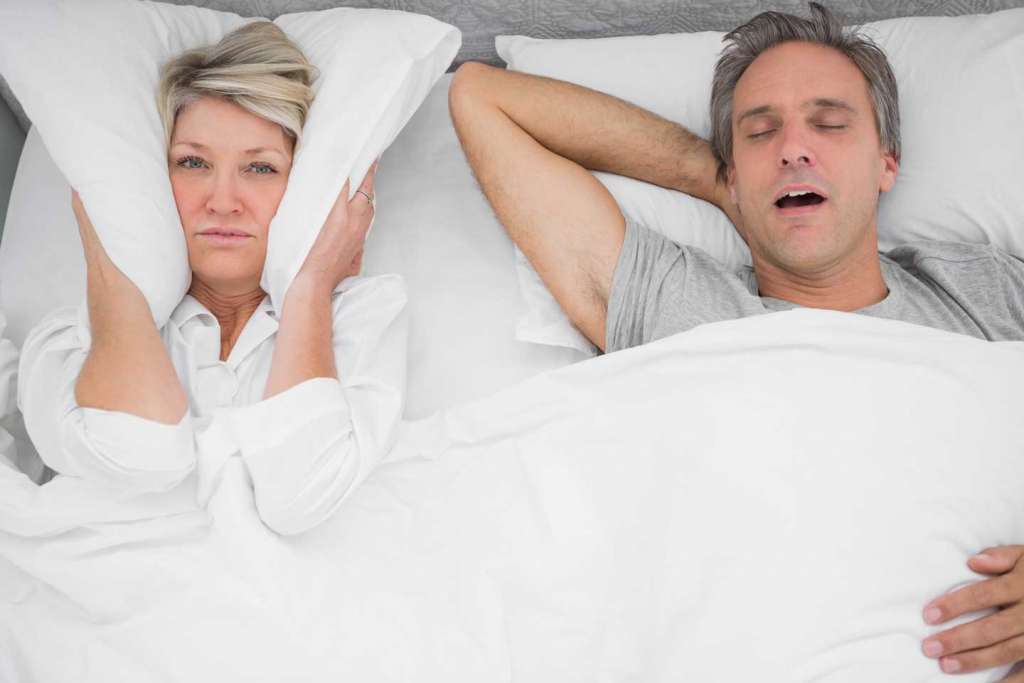 woman covering her ears with pillow to block out man next to her who is snoring.