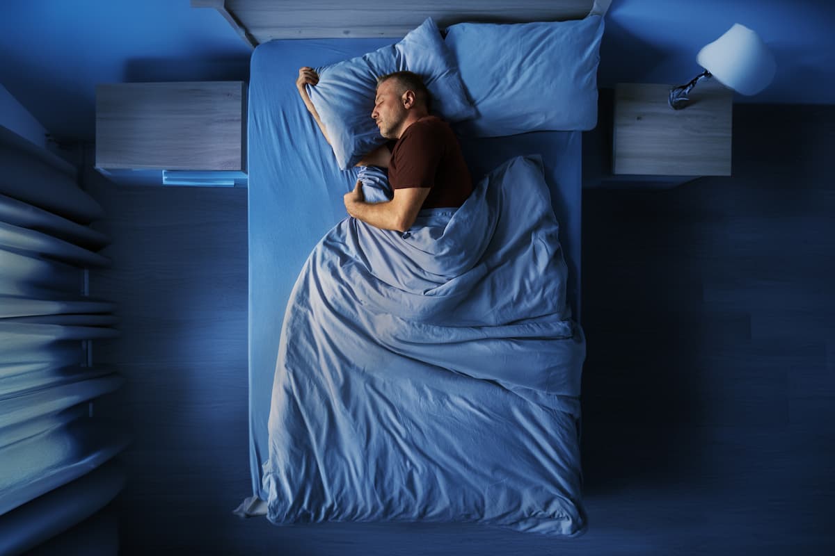 pineal-gland-melatonin-deep-sleep A top-down view of a man sleeping peacefully in a comfortable bed at night, illuminated by cool, dark blue lighting.